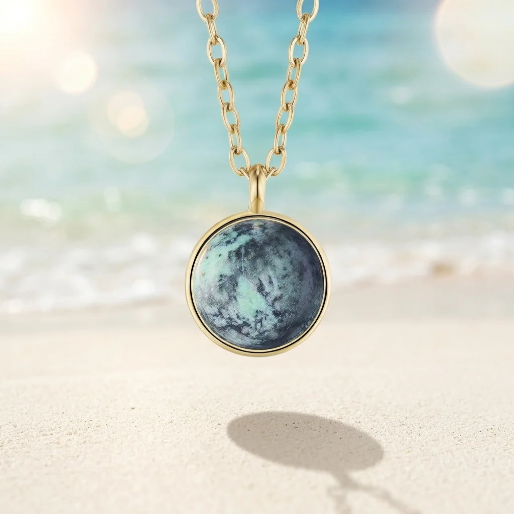Goldkette mit rundem Anhänger, der einen blau-grün marmorierten Stein im Planeten-Design zeigt. Die Kette schwebt vor einem verschwommenen Hintergrund aus hellem Sandstrand und türkisblauem Meer.