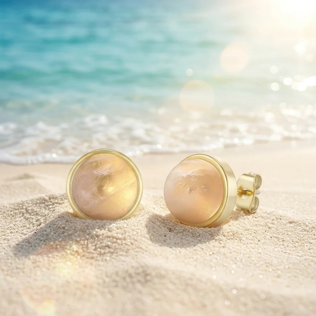 Zwei goldene Ohrstecker mit runden, pfirsichfarbenen, schimmernden Edelsteinen liegen auf hellem Sandstrand. Im Hintergrund ist blau-türkises Meer mit sanften Wellen und Sonnenschein zu sehen. Ein Ohrstecker ist frontal ausgerichtet, der andere zeigt leicht den Ohrsteckerverschluss.