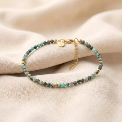 Nahaufnahme eines filigranen Armbands aus facettierten, türkisgrünen Natursteinperlen und kleinen goldenen Kügelchen. Es zeigt einen eleganten Goldverschluss mit Verlängerungskettchen auf einem hellen, drapierten Stoffhintergrund.