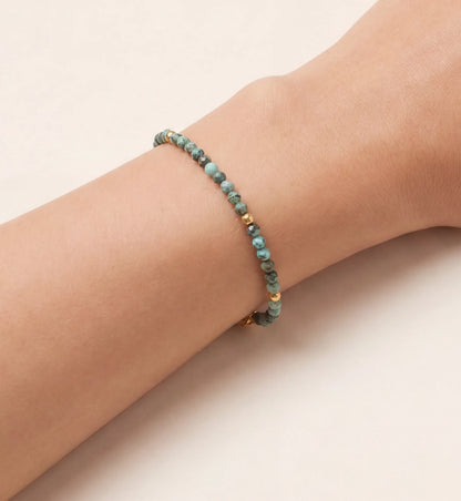 Nahaufnahme eines zarten Armbandes mit facettierten, türkisblauen Edelsteinperlen und glänzenden goldenen Akzentperlen, elegant am weiblichen Handgelenk getragen. Das filigrane Schmuckstück betont die Haut mit seinem natürlichen Charme.