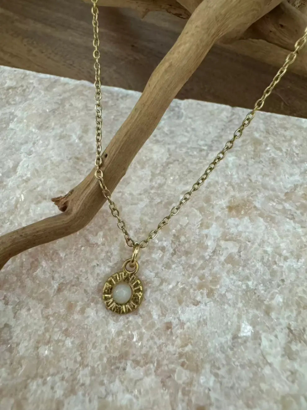 Elegante goldene Kette mit einem runden Anhänger, der einen schimmernden weißen Stein, möglicherweise Opal oder Mondstein, in einer detaillierten Goldfassung zeigt. Präsentiert auf hellem Naturstein mit einem Ast, betont sie minimalistischen, natürlichen Schmuck.