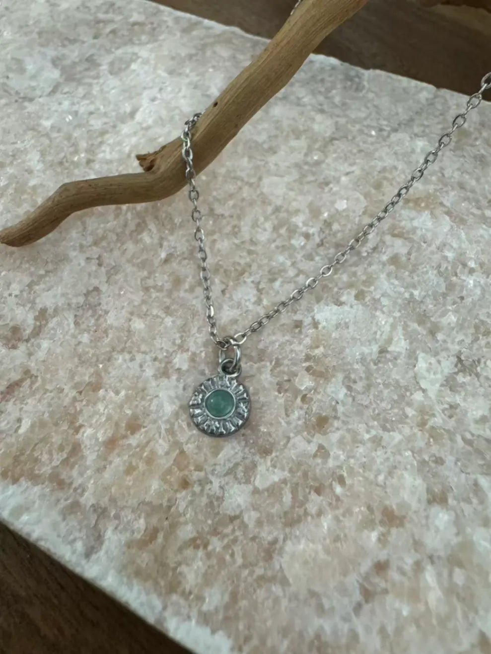 Detailaufnahme einer filigranen Silberkette mit rundem Anhänger. Dieser präsentiert einen zentralen türkisgrünen Edelstein, eingefasst in ein verziertes silbernes Blütenmuster, liegend auf einer hellen Marmoroberfläche mit Treibholz.